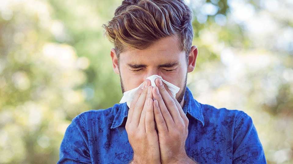 Un hombre con camisa azul sostiene ambas manos frente a su rostro en un entorno al aire libre, con fondo borroso de naturaleza. No se observa texto ni dispositivos auditivos visibles.