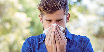 Un hombre con camisa azul está cubriendo su boca con ambas manos en un entorno al aire libre, rodeado de árboles y luz natural. No se identifican audífonos ni texto visible.