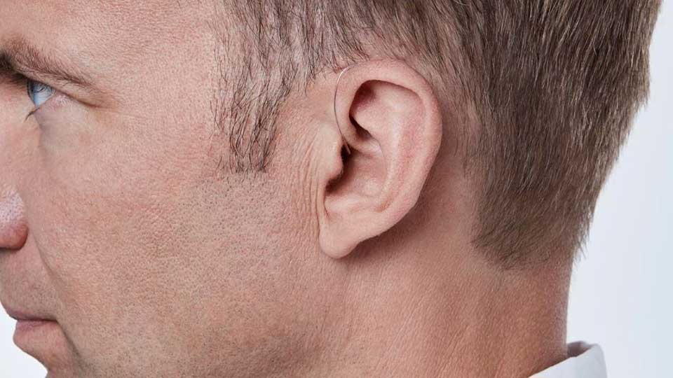 Un oído de un hombre adulto utiliza un audífono discreto detrás de la oreja. Contexto en un entorno bien iluminado, posiblemente una clínica auditiva o ambiente profesional.