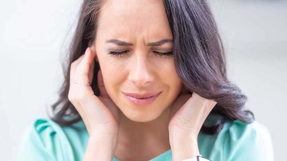 Una persona con cabello oscuro y suelto lleva puestas las manos en ambos costados de su cabeza. Viste una blusa verde en un entorno iluminado y desenfocado.