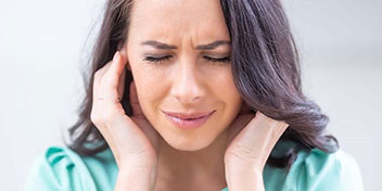 Una mujer con cabello oscuro mantiene ambas manos sobre sus oídos, sugiriendo molestias auditivas. El entorno es claro y minimalista, posiblemente relacionado con una clínica auditiva o consulta de audiología.