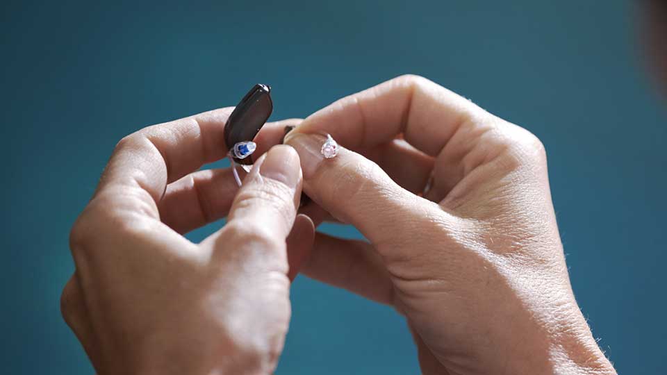 Manos sosteniendo un audífono discreto, posiblemente de la marca Audika, con un fondo azul desenfocado. Ideal para cuidado auditivo en una clínica especializada como Audika o consulta con audiológos.