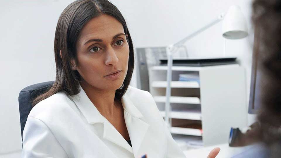 Una audioprotesista con bata blanca gesticula durante una consulta en una clínica auditiva, rodeada de muebles de oficina como estanterías, lámpara y escritorios, fomentando servicios de prueba de audición.