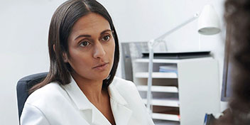 Una audioprotesista vestida con bata blanca escucha atentamente a un paciente en una clínica auditiva, rodeada de muebles blancos, lámpara y archivadores organizados en el fondo. No hay texto visible.