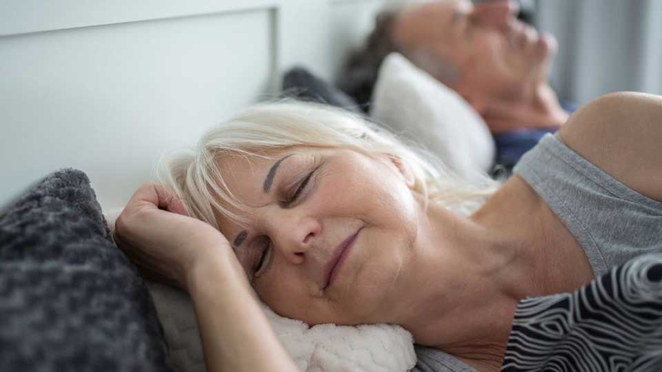 Una persona con cabello blanco descansa sobre un sofá gris en un entorno doméstico, acompañada por otra persona al fondo.