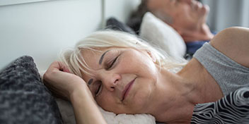 Una pareja de personas mayores descansa en una cama con cojines claros en un entorno hogareño tranquilo. No hay texto ni audífonos visibles en la imagen.