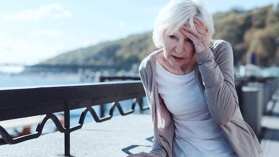 Una mujer mayor, vestida con ropa clara y un cárdigan, se sienta al aire libre apoyada sobre un barandal metálico, con una mano cubriendo parcialmente su rostro; el entorno incluye un paisaje costero con colinas y agua bajo un cielo despejado. No hay texto visible en la imagen.