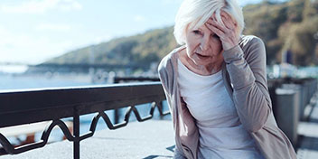 Una mujer mayor de cabello blanco, vestida con ropa clara, se lleva la mano al rostro. Está sentada junto a una barandilla en un entorno exterior soleado, cerca del agua.
