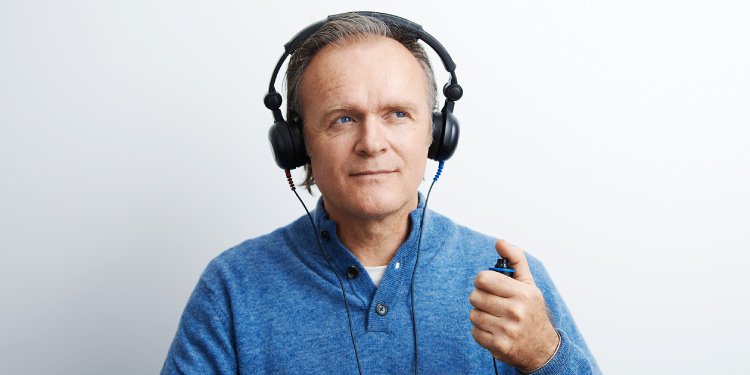 Un hombre mayor, con audífonos colocados, sostiene un pulsador manual mientras realiza una prueba de audición en un audiólogo o clínica auditiva. Fondo blanco y ropa casual azul.