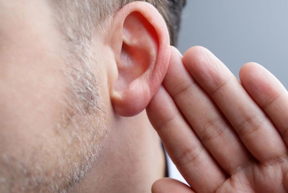 Una oreja masculina en primer plano, con una mano acercándose en gesto de escuchar. Fondo gris neutro, ideal para ilustrar temas de pérdida auditiva, audífonos o consultas en clínicas auditivas.