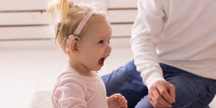 Una niña pequeña lleva un audífono con una diadema rosa, probablemente un dispositivo para pérdida auditiva, en un ambiente interior iluminado. A su lado, un adulto interactúa con ella.