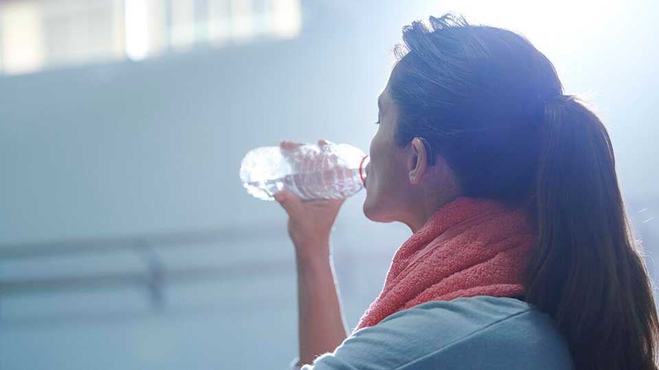 Una mujer con cabello atado bebe agua de una botella transparente mientras lleva una toalla rosada alrededor del cuello en un entorno iluminado, posiblemente un gimnasio con fondo desenfocado.