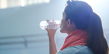 Una mujer, con una toalla naranja en el cuello, bebe agua de una botella en un ambiente iluminado que parece un gimnasio, destacando actividad física y cuidados personales.