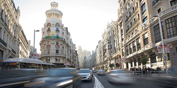 Una bulliciosa avenida urbana con coches en movimiento domina la escena; edificios clásicos alinean ambos lados, destacando uno con cúpula blanca. Contexto diurno en ciudad moderna, posiblemente Madrid.