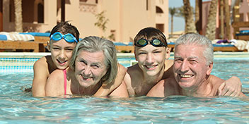 Cuatro personas nadan juntas en una piscina, disfrutando de un día soleado. Al fondo, se observan edificios de estilo mediterráneo, tumbonas y palmeras en un ambiente cálido.