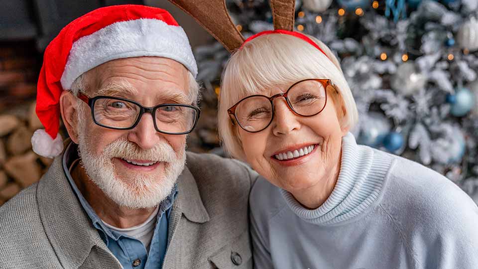 Dos personas mayores, una con gorro de Papá Noel y otra con diadema de reno, posan juntas frente a un árbol de Navidad decorado con luces y adornos festivos.