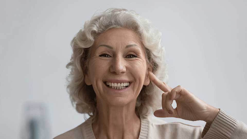 Una persona mayor con cabello canoso apunta a su oído con un dedo, en un entorno interior minimalista. El contexto sugiere un enfoque en salud auditiva o audífonos.