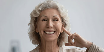 Una persona mayor con cabello canoso señala su oído con el dedo índice, sugiriendo problemas de audición. Fondo borroso y claro, posiblemente en una clínica auditiva o entorno neutro.