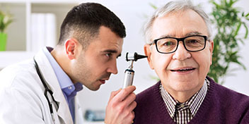 Un audiólogo usa un otoscopio para examinar el oído de un hombre mayor en una clínica auditiva luminosa, rodeada de muebles blancos y decoraciones con plantas verdes. No hay texto visible.