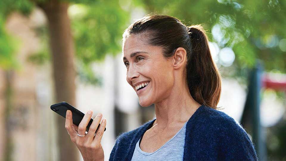 Una mujer sostiene un smartphone cerca de su boca, aparentemente usando la función de voz. Lleva el cabello recogido en una coleta y está al aire libre, rodeada de árboles.