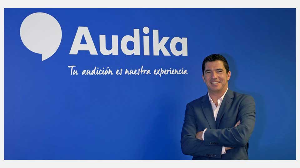Un hombre de traje con los brazos cruzados frente a una pared azul con el logotipo de Audika y su lema: ’Tu audición es nuestra experiencia’. Contexto profesional y corporativo.