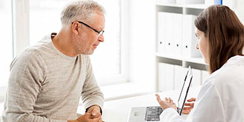 Un hombre mayor, sentado frente a una audioprotesista, revisa información en una computadora en un consultorio iluminado con estanterías y ventanas, posiblemente durante una consulta para audífonos o prueba auditiva.