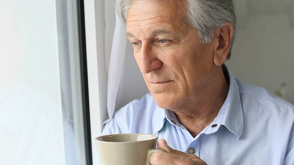 Un hombre de cabello canoso sostiene una taza de cerámica beige cerca de una ventana, vistiendo una camisa azul claro. El entorno parece ser un espacio tranquilo y luminoso.