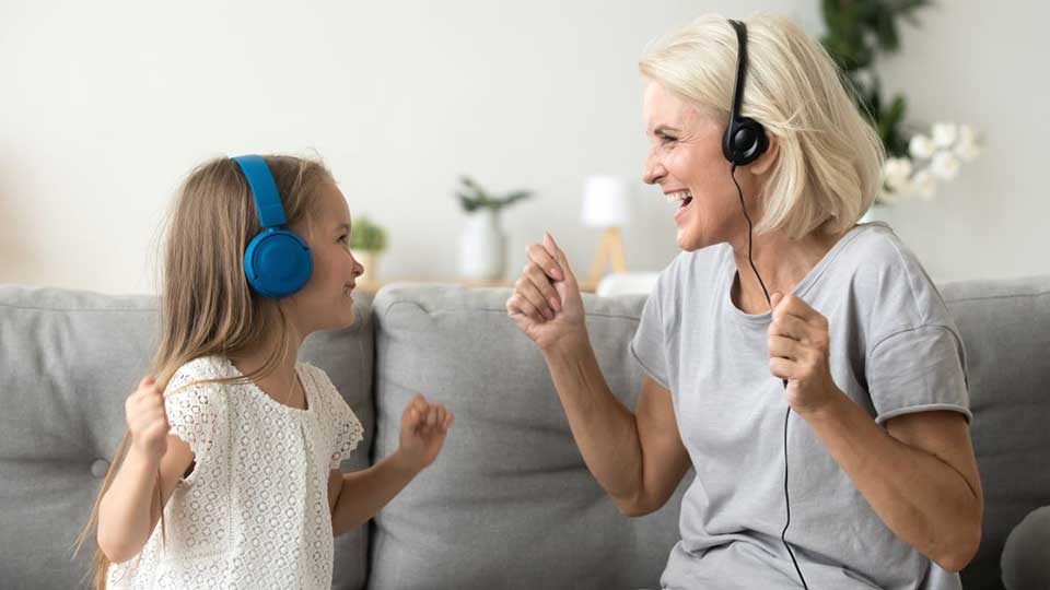 Una mujer mayor y una niña usan auriculares mientras bailan sonrientes en un sofá gris. El entorno es una sala luminosa con decoración minimalista, incluyendo plantas y lámparas.