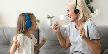 Una niña con auriculares azules y una mujer mayor con auriculares negros sonríen y gesticulan mientras están sentadas en un sofá gris en un ambiente hogareño iluminado y decorado con plantas.