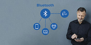 Un hombre de pie sostiene un dispositivo móvil. En el contexto, un gráfico destaca conexiones Bluetooth con íconos que representan smartphone, música, campana de alerta y televisión, sobre un fondo azul.