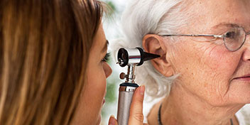 Un audiólogo examina el oído de una persona mayor con un otoscopio en una clínica auditiva. El entorno es claro, resaltando el enfoque en el cuidado auditivo y las pruebas auditivas.