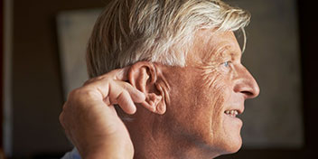 Un hombre mayor toca su oído con la mano en un ambiente interior, posiblemente relacionado con el uso o revisión de audífonos en un contexto de cuidado auditivo. No se observa texto visible.