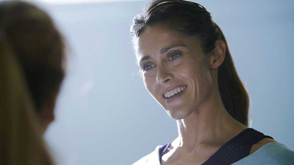 Una mujer de cabello recogido lleva ropa deportiva y está frente a otra persona, en un ambiente iluminado con luz suave y tonalidades azules. No hay texto visible.