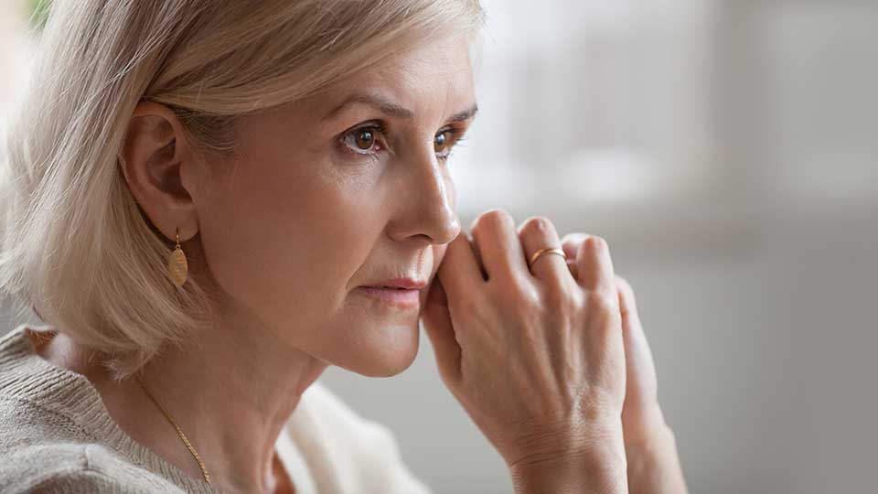 Una mujer mayor, con cabello rubio corto y un pendiente dorado, sostiene sus manos cerca de su rostro en un ambiente iluminado, posiblemente relacionada con cuidado auditivo o audífonos.