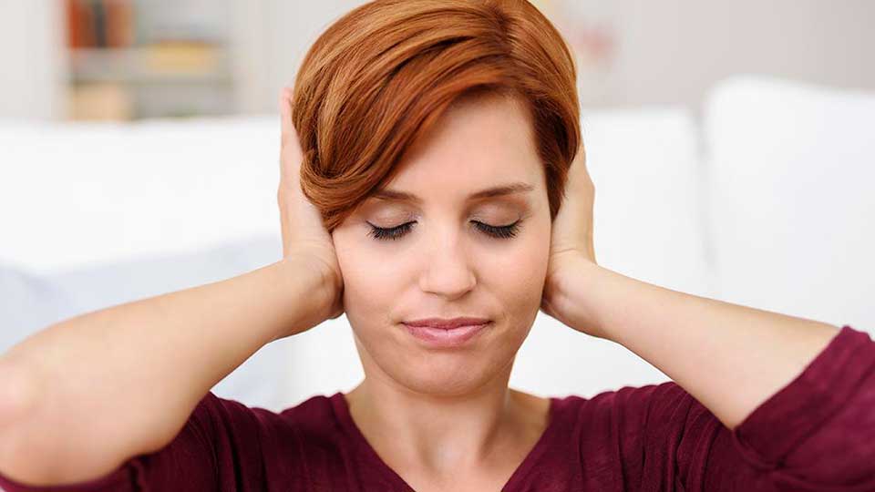 Persona con cabello rojizo cubriendo sus oídos con las manos, posiblemente reflejando incomodidad auditiva; en un entorno interior con fondo desenfocado, sugestivo de una sala residencial.