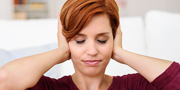 Una persona con cabello rojizo se cubre los oídos con ambas manos, transmitiendo incomodidad o sensibilidad auditiva, en un entorno hogareño con fondo desenfocado, posiblemente relacionado con problemas de audición.