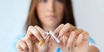 Una persona sostiene y rompe un cigarrillo en sus manos, representando una acción de dejar de fumar. Fondo difuminado sugiere un ambiente interior o clínico. No hay texto visible.