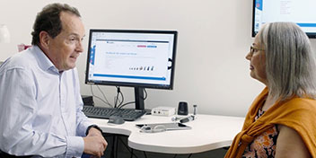 Un audioprotesista conversa con una mujer mayor en una clínica auditiva. En el fondo, una pantalla muestra gráficos relacionados con evaluaciones auditivas. Sobre el escritorio, herramientas para pruebas de audición.