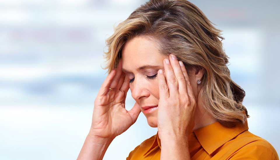 Una mujer con cabello rubio está tocándose ambas orejas con las manos. Lleva una blusa anaranjada. El fondo es claro y desenfocado, evocando un entorno clínico o profesional.