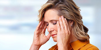 Una mujer con cabello rubio lleva un audífono discreto en el oído derecho, mientras se toca las sienes con ambas manos. Fondo desenfocado en tonos claros.