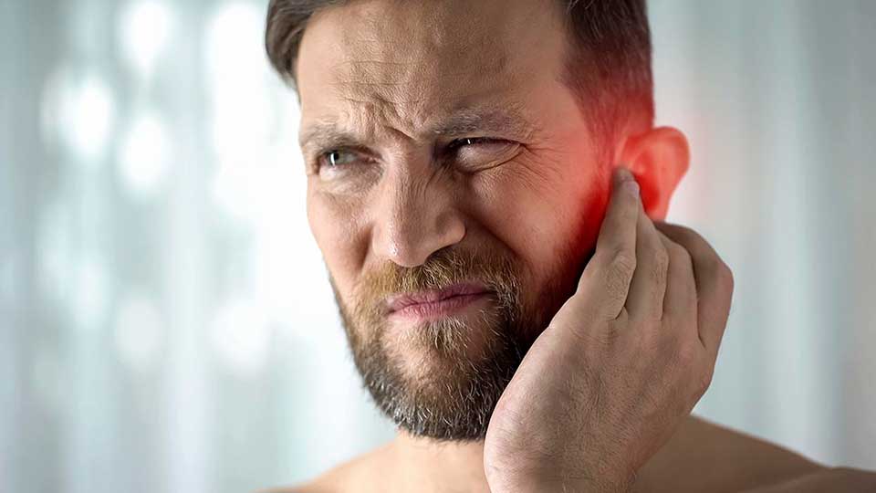 Un hombre adulto se toca el oído con la mano, posiblemente sintiendo molestias auditivas, en un entorno interior despejado con iluminación suave. No se observan audífonos ni texto visible.