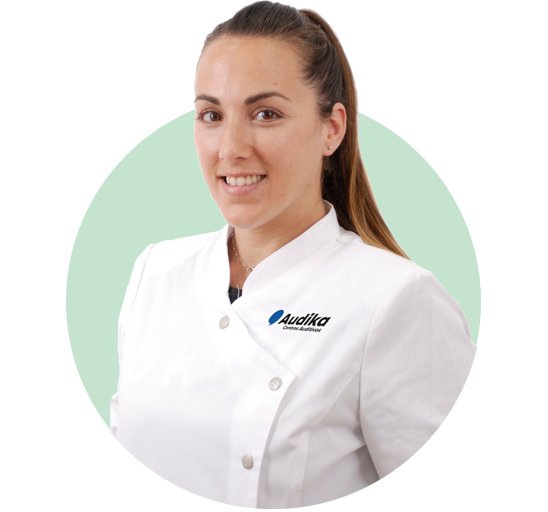 Una profesional de la salud auditiva viste un uniforme blanco con el logotipo de ’Audika Cuidamos tu audición’ en el pecho. Fondo circular verde claro.