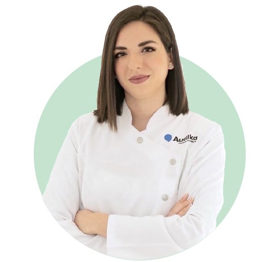 Una audióloga con bata blanca que lleva el logo de Audika, brazos cruzados, en un fondo circular verde claro. Promueve servicios de audición como pruebas auditivas y audífonos especializados.