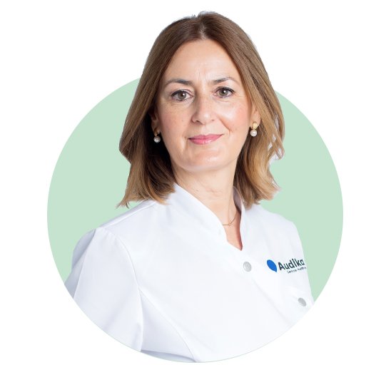 Una profesional de la salud auditiva, con bata blanca del logo de Audika, está sobre un fondo verde claro. Su cabello es rubio y lleva aretes discretos. Texto visible: ’Audika Hearing Care Experts’.
