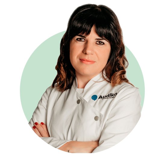 Una audioprotesista con bata blanca cruzada de botones, brazos cruzados y cabello ondulado. El logo de Audika, especializado en cuidado auditivo, se encuentra en su uniforme. Fondo circular verde claro.