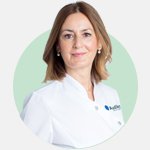 Una profesional de la salud auditiva con bata blanca que lleva el logotipo de Audika realiza su labor dentro de un entorno con fondo verde claro, representando servicios de audiología y cuidado auditivo.