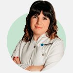 Una profesional con bata blanca de Audika, brazos cruzados, en un fondo verde claro. Su rostro está difuminado. El logo de Audika se distingue en su uniforme.