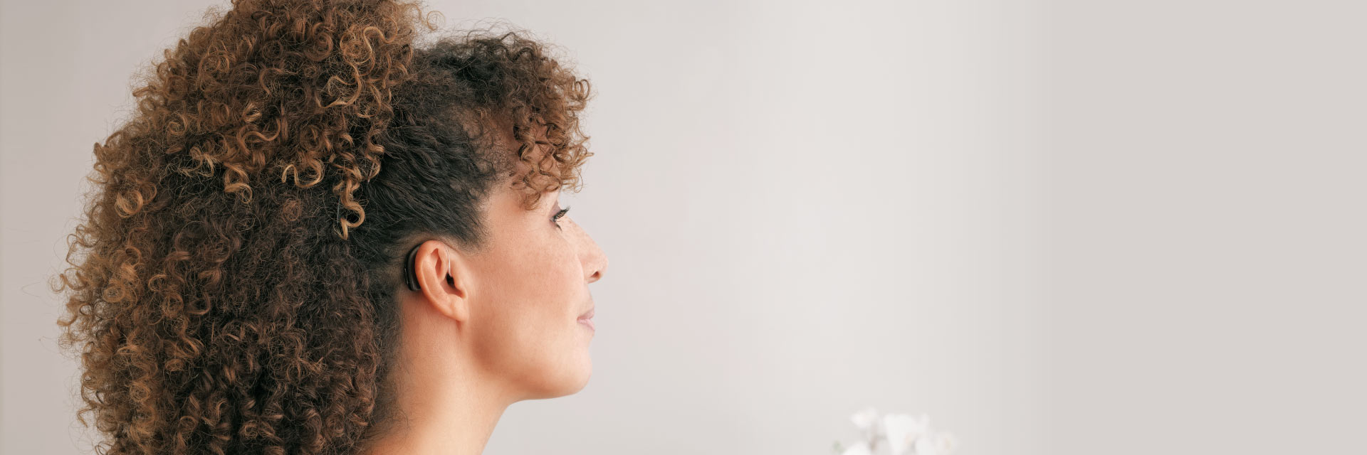 Un audífono detrás del oído de una persona con cabello rizado oscuro y cobrizo. La escena se desarrolla en un entorno minimalista con fondo claro y flores blancas desenfocadas.