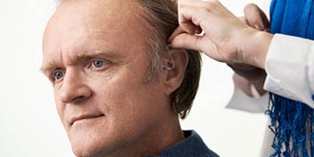 Una audioprotesista ajusta un audífono detrás de la oreja de una persona en un entorno clínico, posiblemente relacionado con Audika, especialista en cuidado auditivo. El fondo es blanco y limpio.