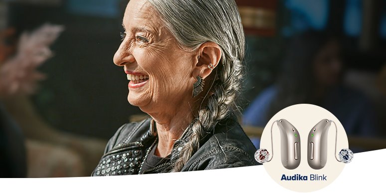 Una persona de cabello gris trenzado usa un audífono discreto detrás de la oreja izquierda. En el contexto, se observa un fondo borroso y el logotipo 'Audika Blink' junto a dos audífonos modernos plateados.